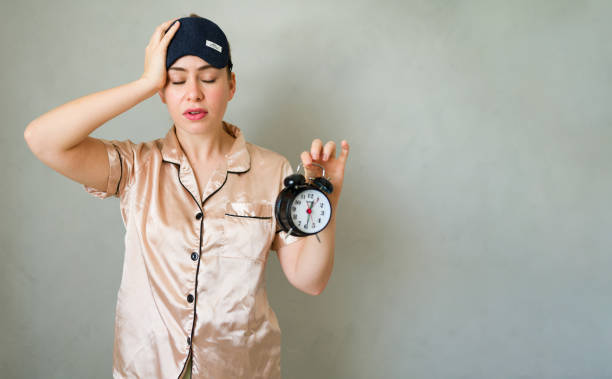 Woman wearing pajamas and an eye mask is having trouble waking up and holding an alarm clock