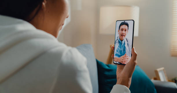 Virtual Reality in Therapy - Sick young Asian woman using phone talk to doctor sitting on sofa in living room at home. Telemedicine and Healthcare concept.